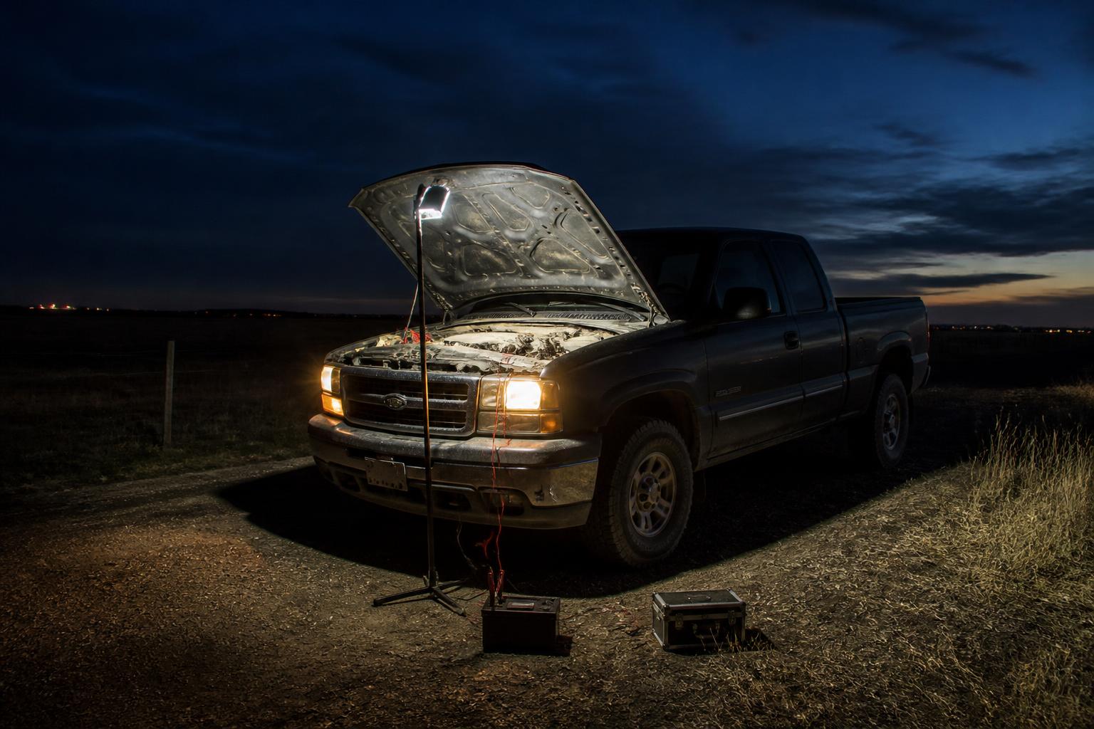 Pickup truck on a rural roadside at dusk with the hood up, lit by a portable LED work light beside a toolbox and battery pack